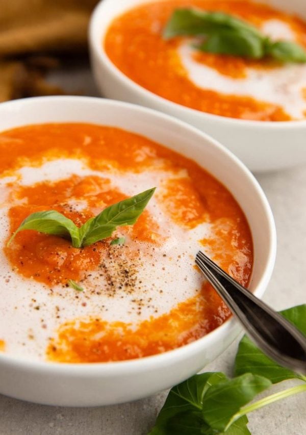 Two bowls of creamy tomato soup with a spoon and fresh basil leaves.