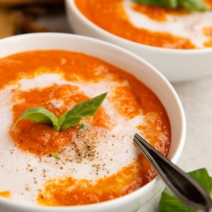 Two bowls of creamy tomato soup with a spoon and fresh basil leaves.