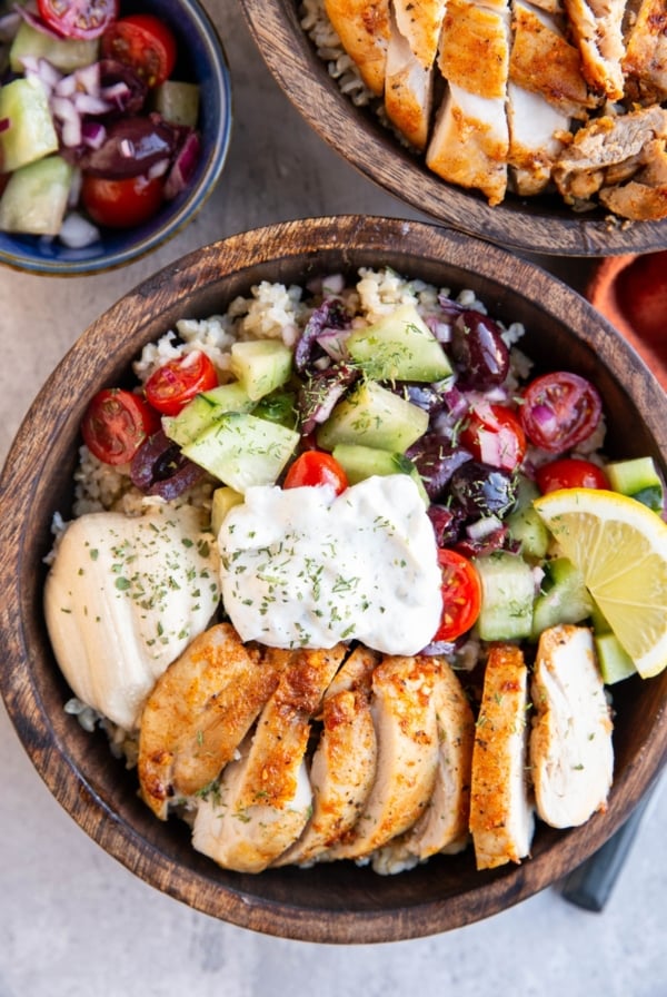 Two wooden bowls full of chicken , hummus, tzatziki sauce, and cucumber tomato salad, ready to serve.