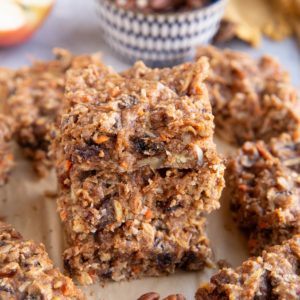 Stack of carrot cake breakfast bars on a sheet of parchment paper with the pecans in the background.