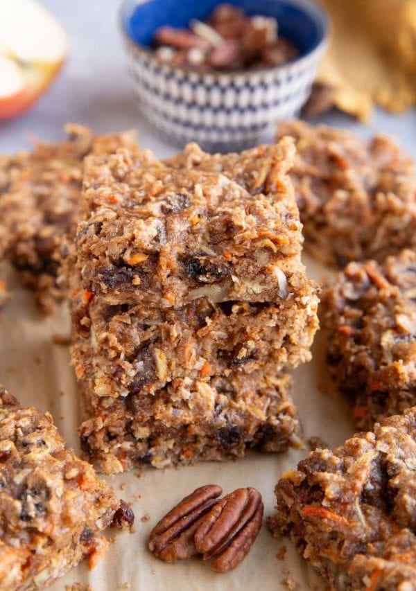 Stack of carrot cake breakfast bars on a sheet of parchment paper with the pecans in the background.