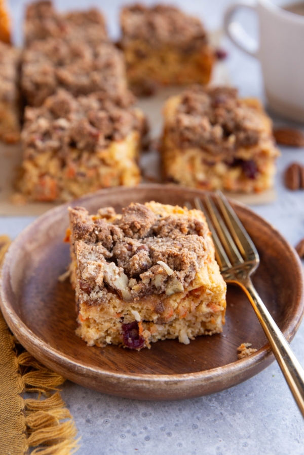 Carrot Cake Coffee Cake on a wooden plate with a golden fork and the rest of the cake in the background.