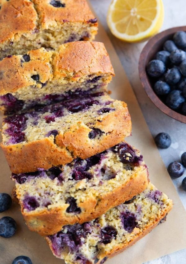 Loaf of lemon blueberry bread cut into slices with a bowl of blueberries and cut lemon in the background.