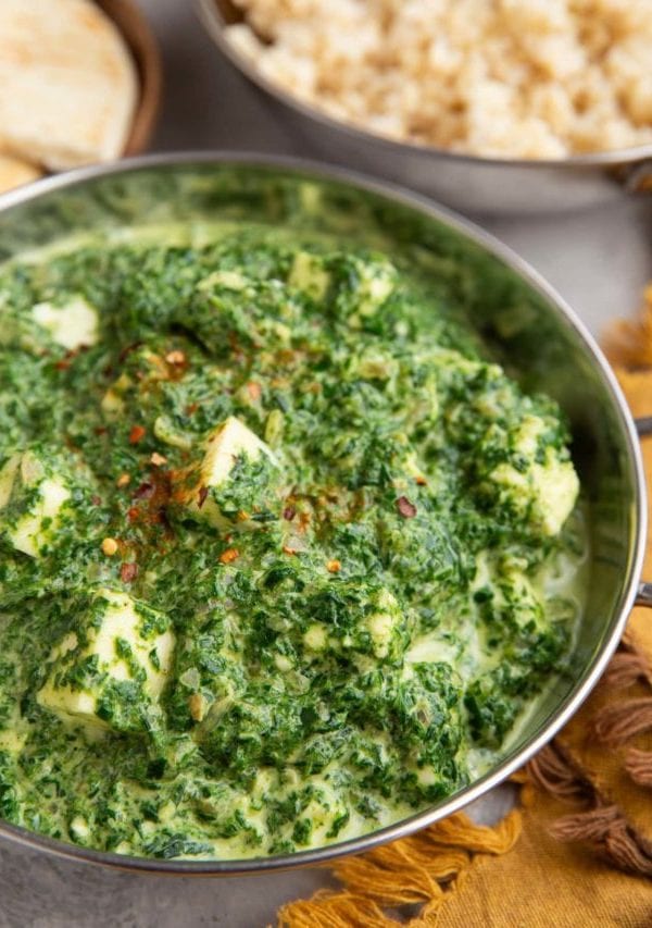 Indian dish full of saag paneer with a dish of brown rice in the background.