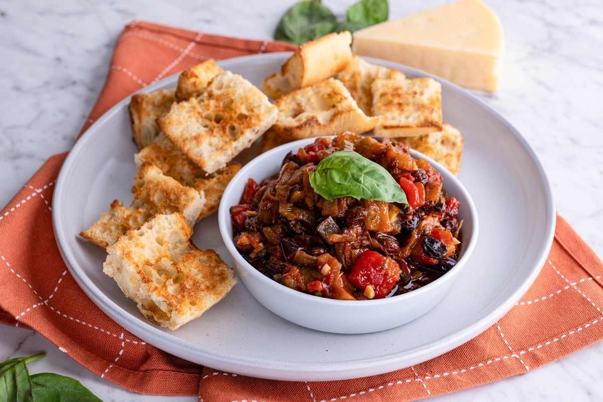 Bowl of eggplant caponata with toasted bread on a plate and parmesan cheese in the background.