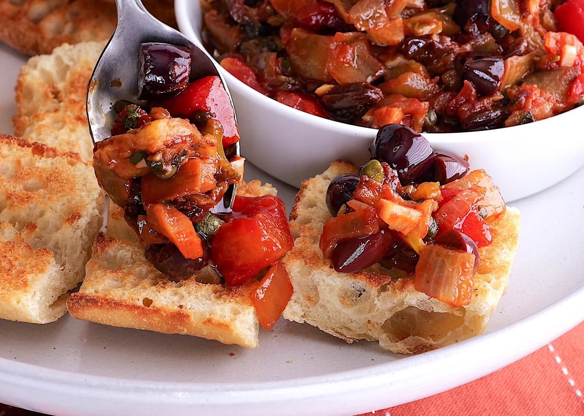 Spoon scooping caponata onto toasted bread.