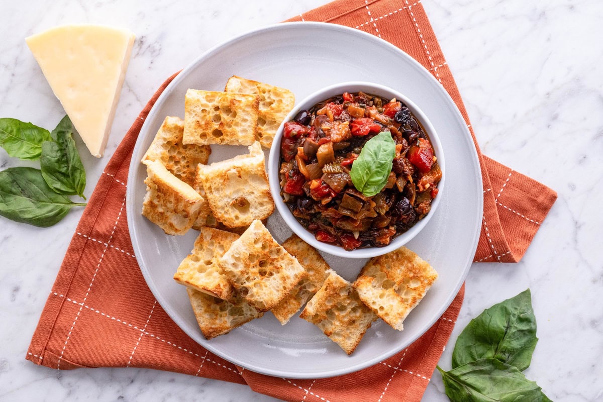 Plate of toasted bread with a bowl of eggplant caponata and fresh parmesan cheese and basil to the side.