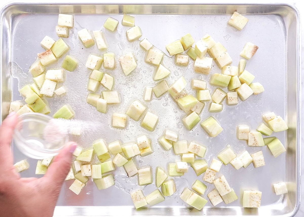 Roasting pan with chopped eggplant cubes tossed in oil, being sprinkled with sea salt.