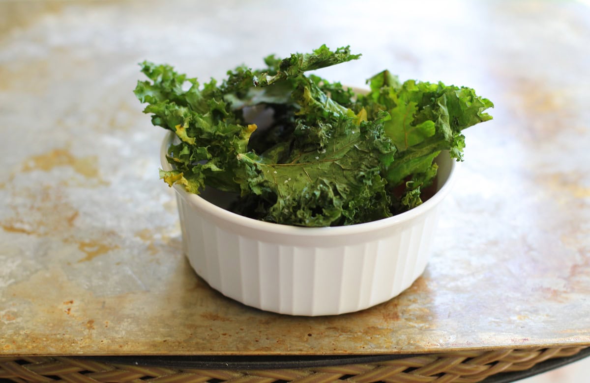 kale chips in a white bowl on top of a baking sheet, fresh out of the oven and ready to snack on.