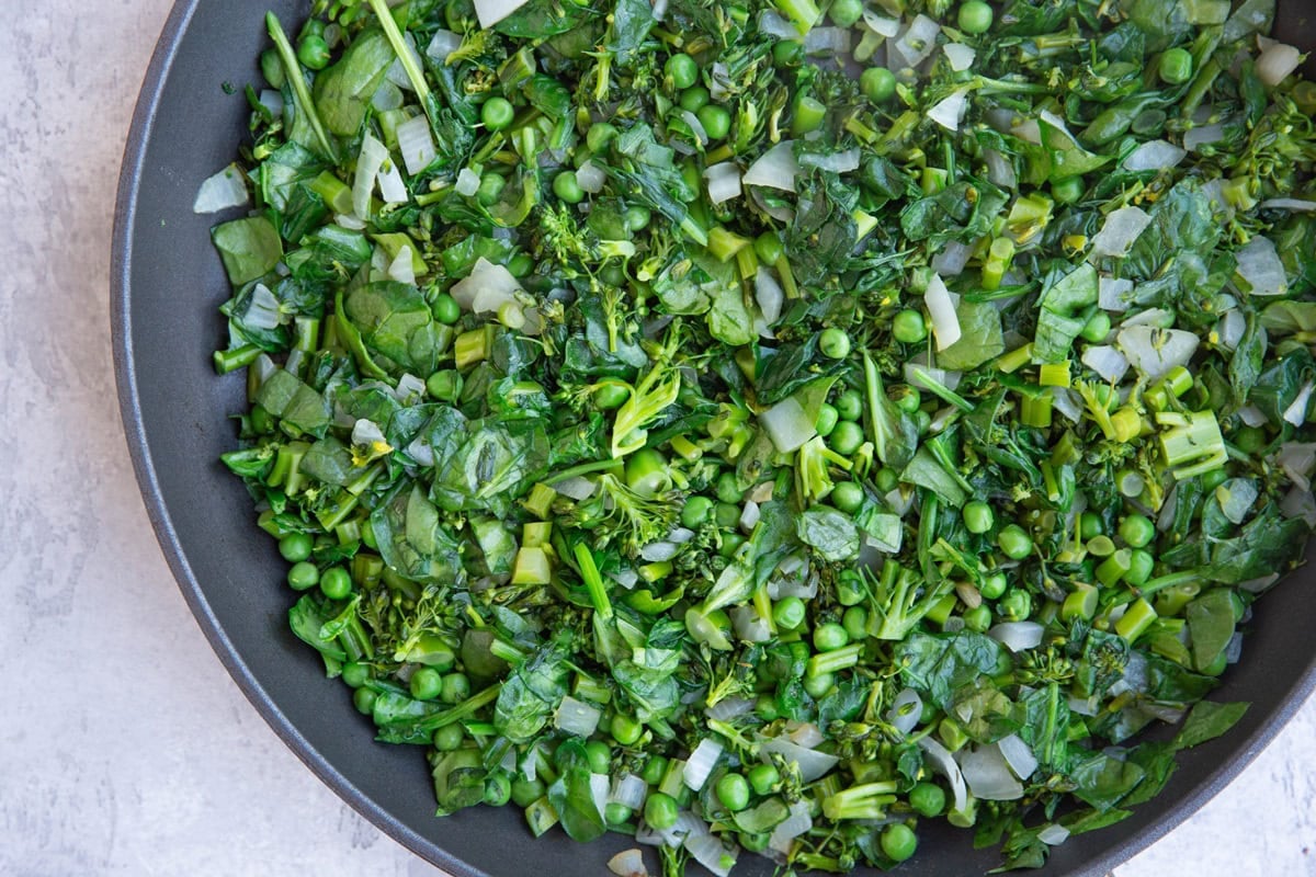 Skillet full of spinach, peas, broccoli, and onions, cooking.