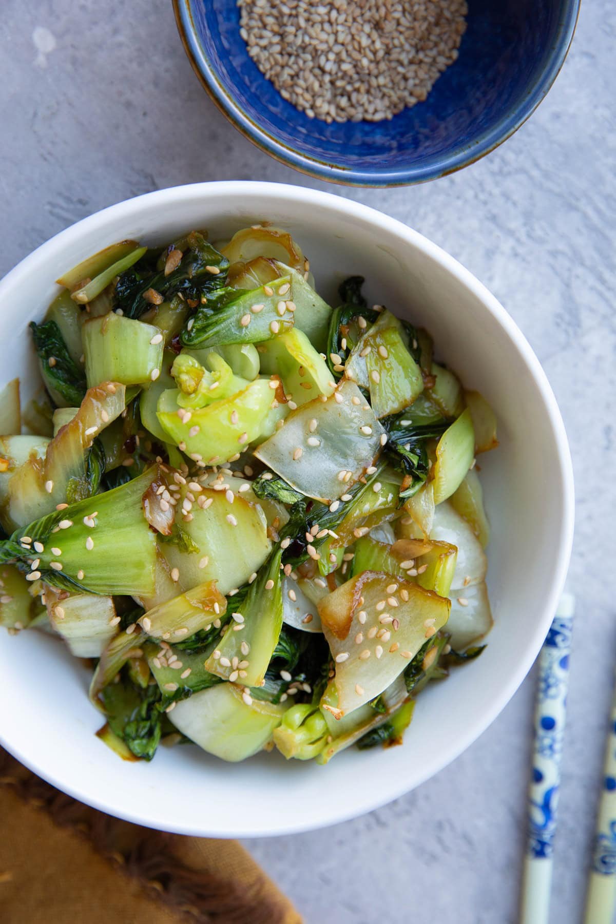 Bowl full of sauteed bok choy with chop sticks to the side and a bowl of sesame seeds.