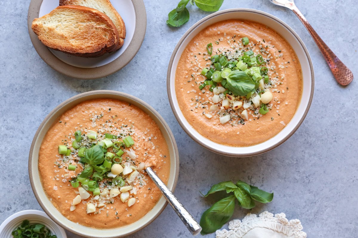 Two bowls of creamy tomato soup with chopped green onions, nuts and seeds sprinkled on top.