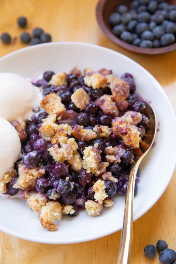 Large bowl full of blueberry crumble with two scoops of vanilla ice cream and a bowl of fresh blueberries to the side.