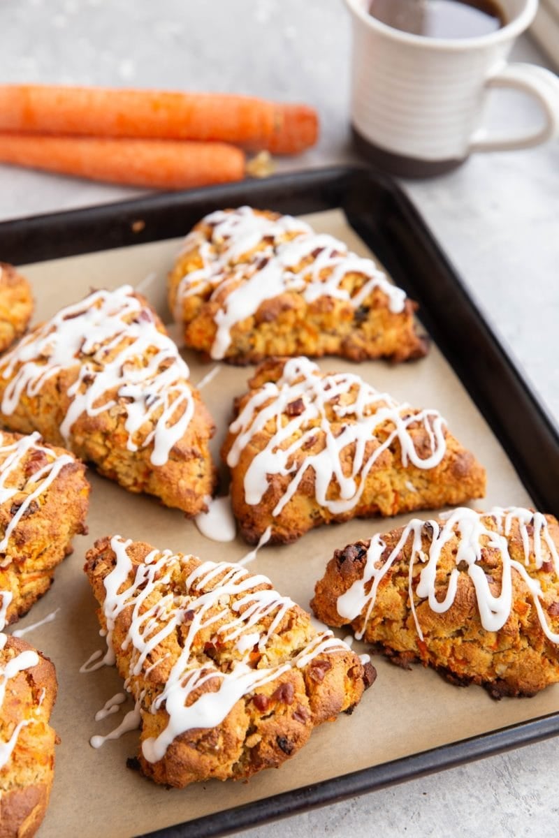 Baking sheet with carrot scones drizzled with glaze with fresh carrots and a mug of coffee in the background.