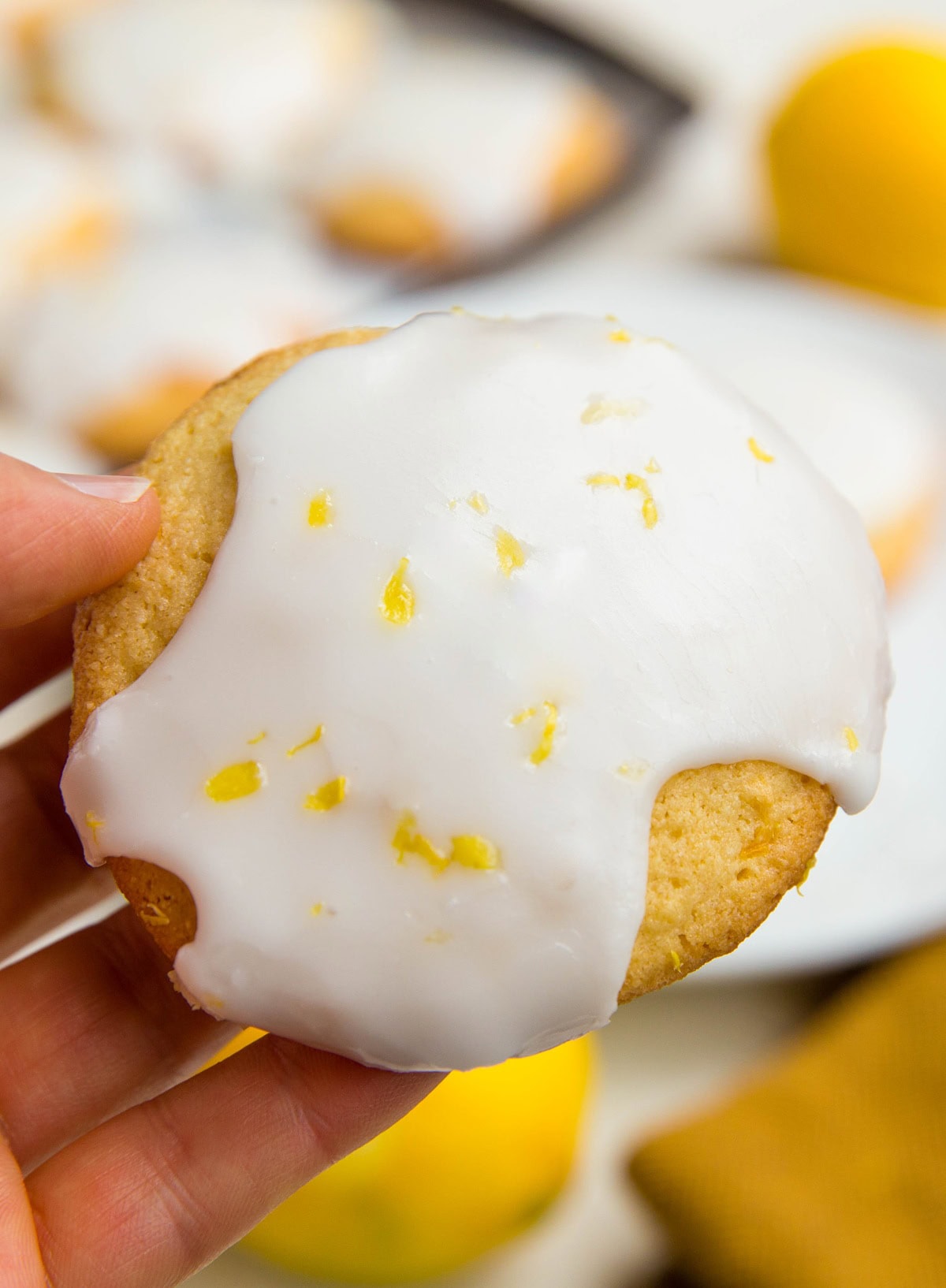Hand holding a glazed lemon cookie with lemon zest on the glaze, ready to eat the cookie.
