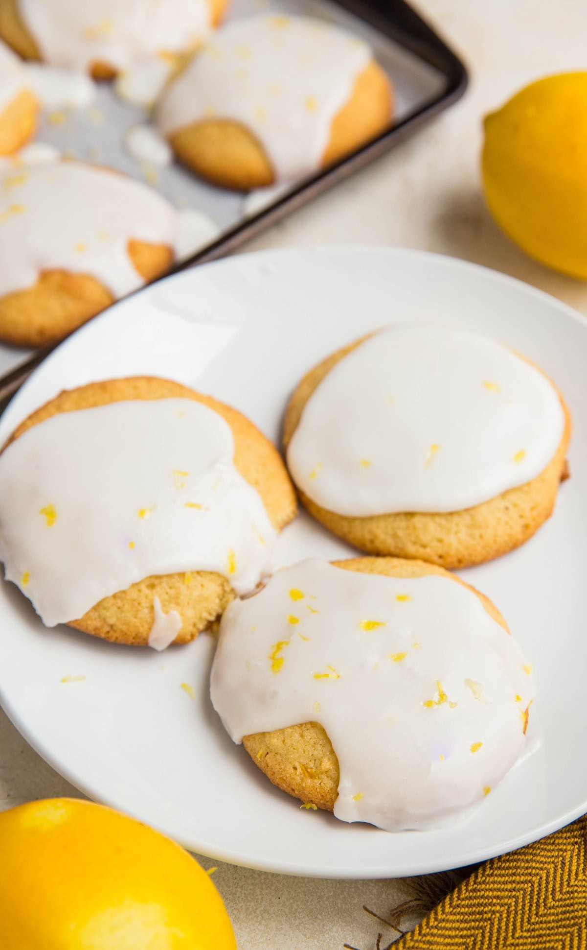 White plate of iced lemon cookies with a cookie sheet of cookies in the background.