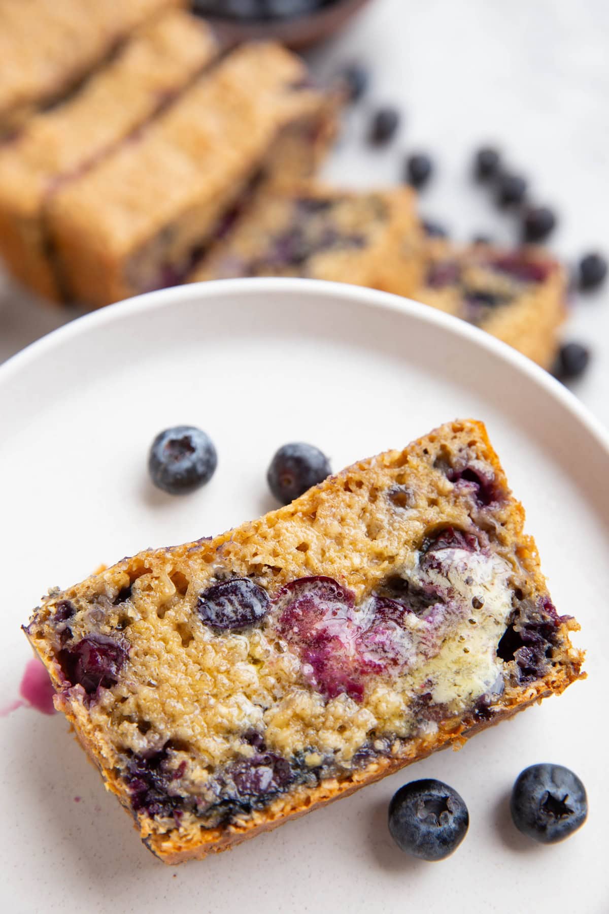 Slice of Blueberry Cottage Cheese Bread with melted butter, ready to eat. The rest of the sliced loaf in the background.