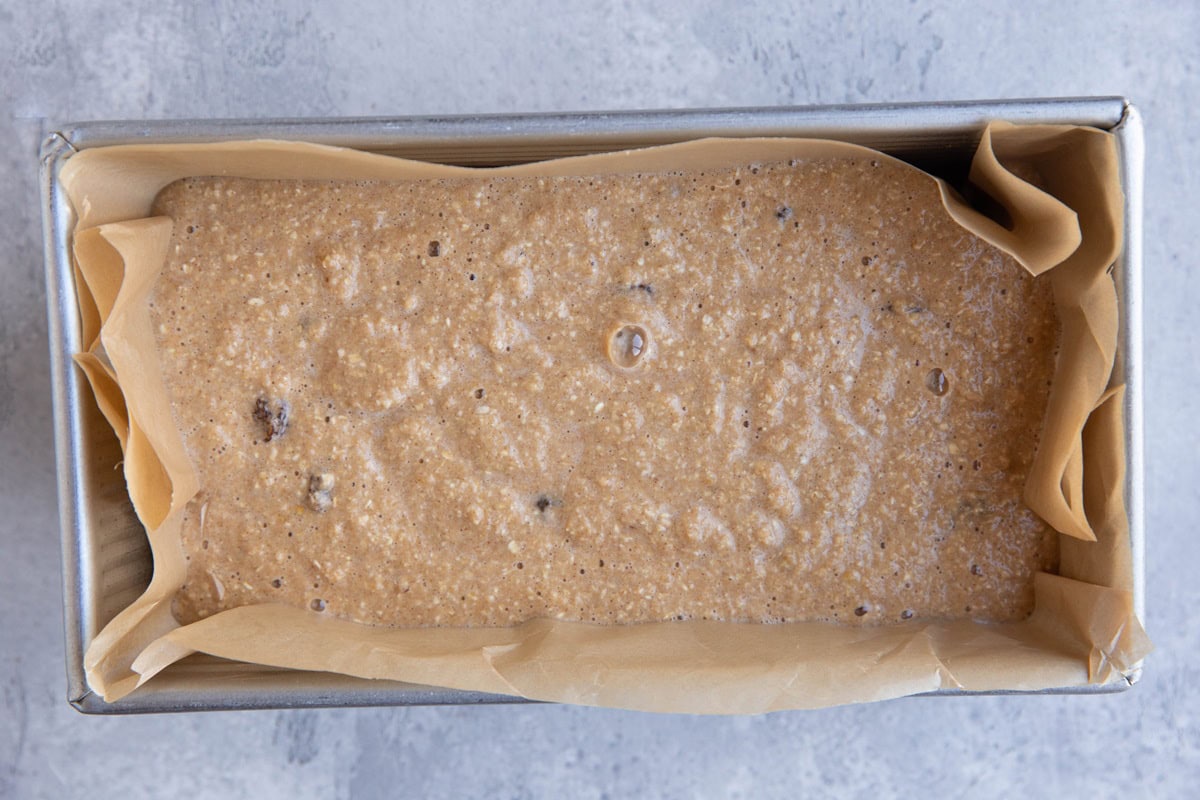 Loaf pan lined with parchment paper with oatmeal raisin bread batter inside.