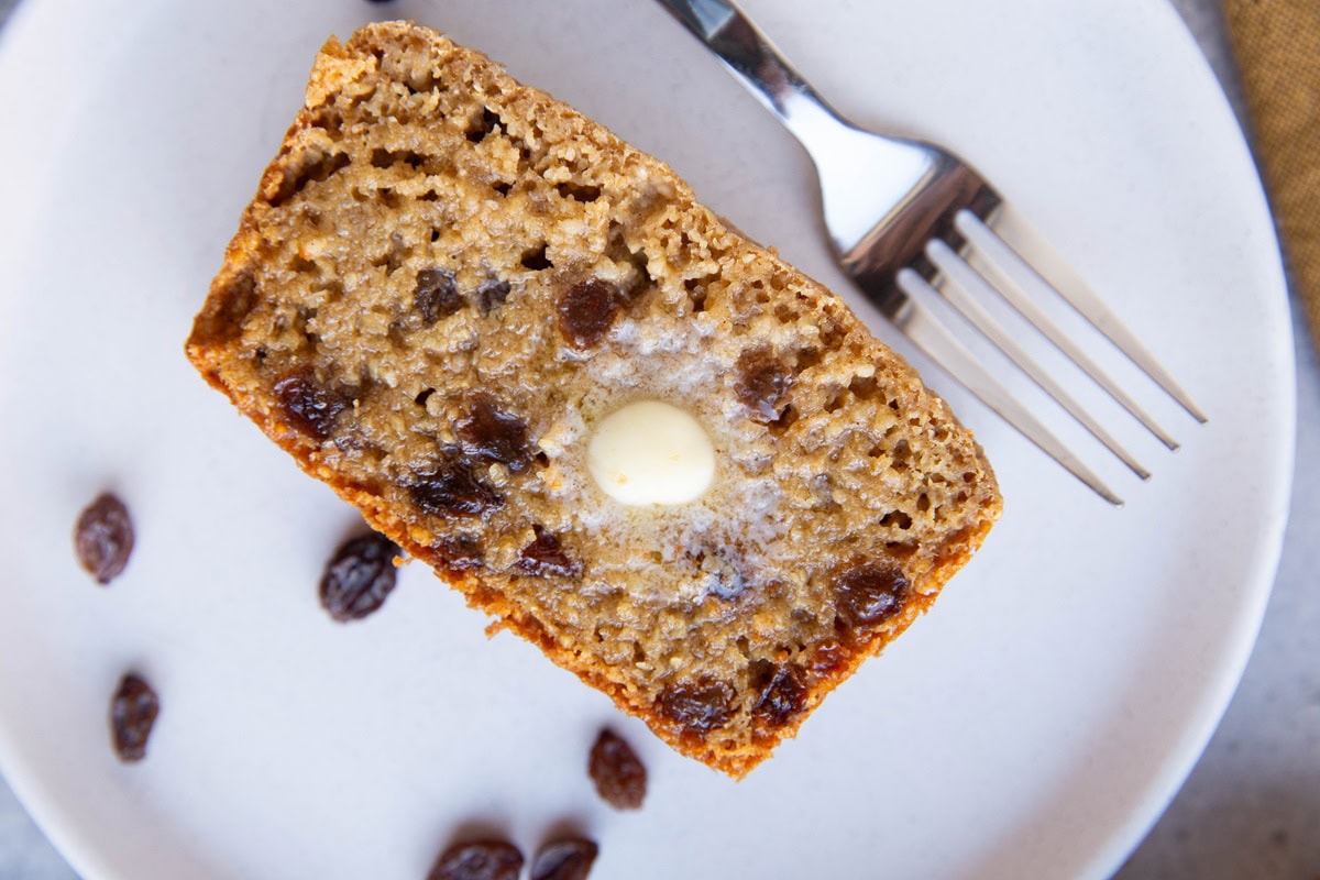 Slice of cinnamon raisin bread on a white plate with raisins to the side.