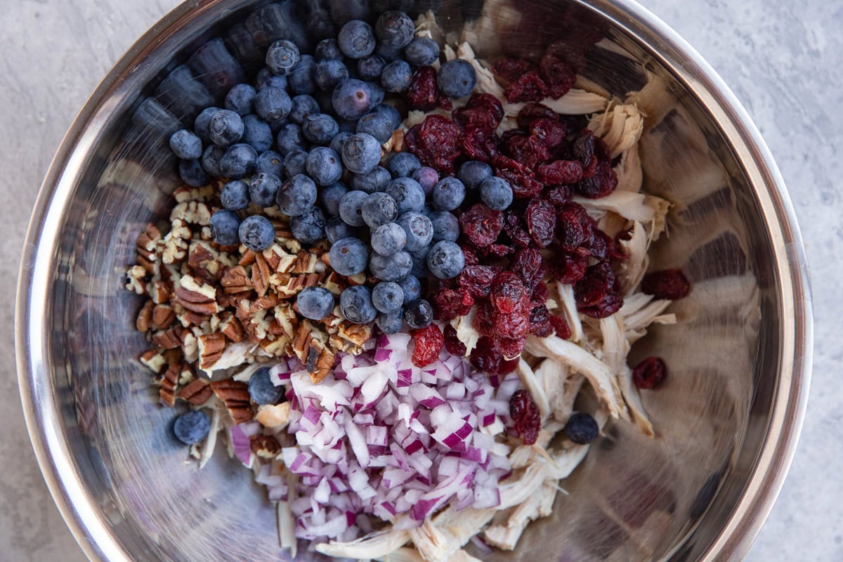 Stainless steel bowl full of shredded chicken, blueberries, dried cranberries, pecans, and onions.