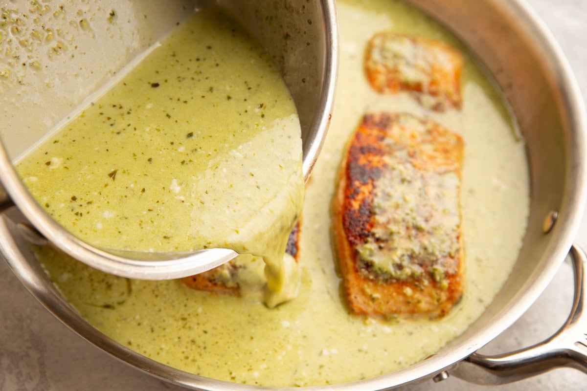 Pouring creamy pesto sauce into a skillet with the salmon.