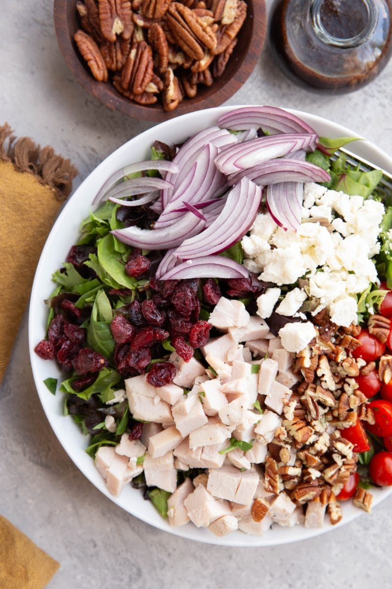 Salad in a large bowl with chicken, pecans, feta cheese, red onion, dried cranberries, and tomatoes