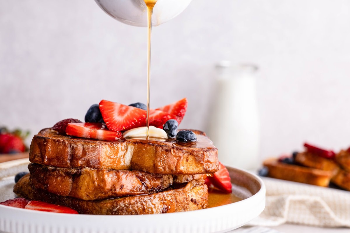 Pouring pure maple syrup over a stack of French toast with berries and butter on top.