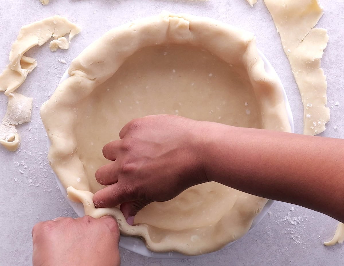 Hands crimping the edges of pie dough in a pie pan.