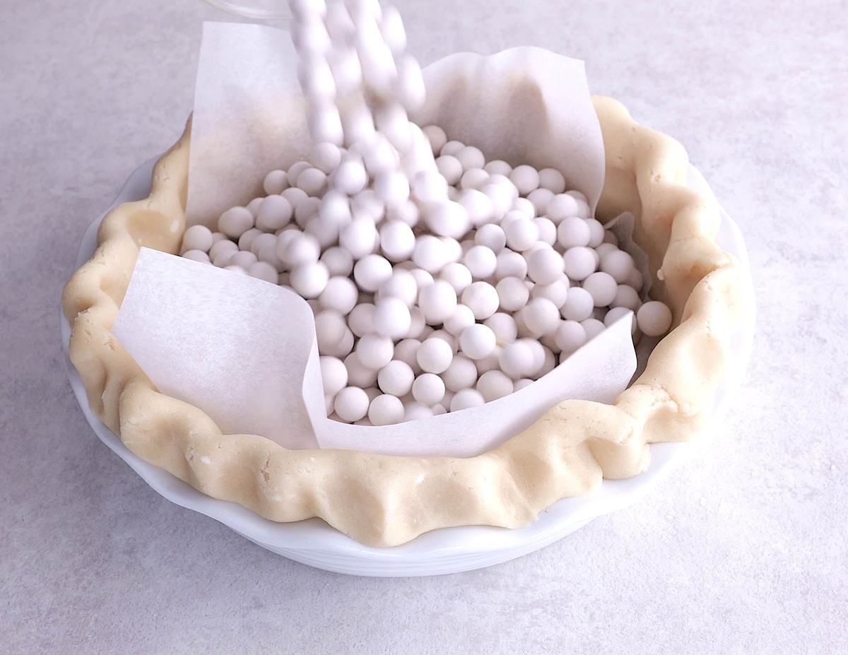 Pie dough in a pie pan covered with parchment paper with pie weights being poured into the pie pan.
