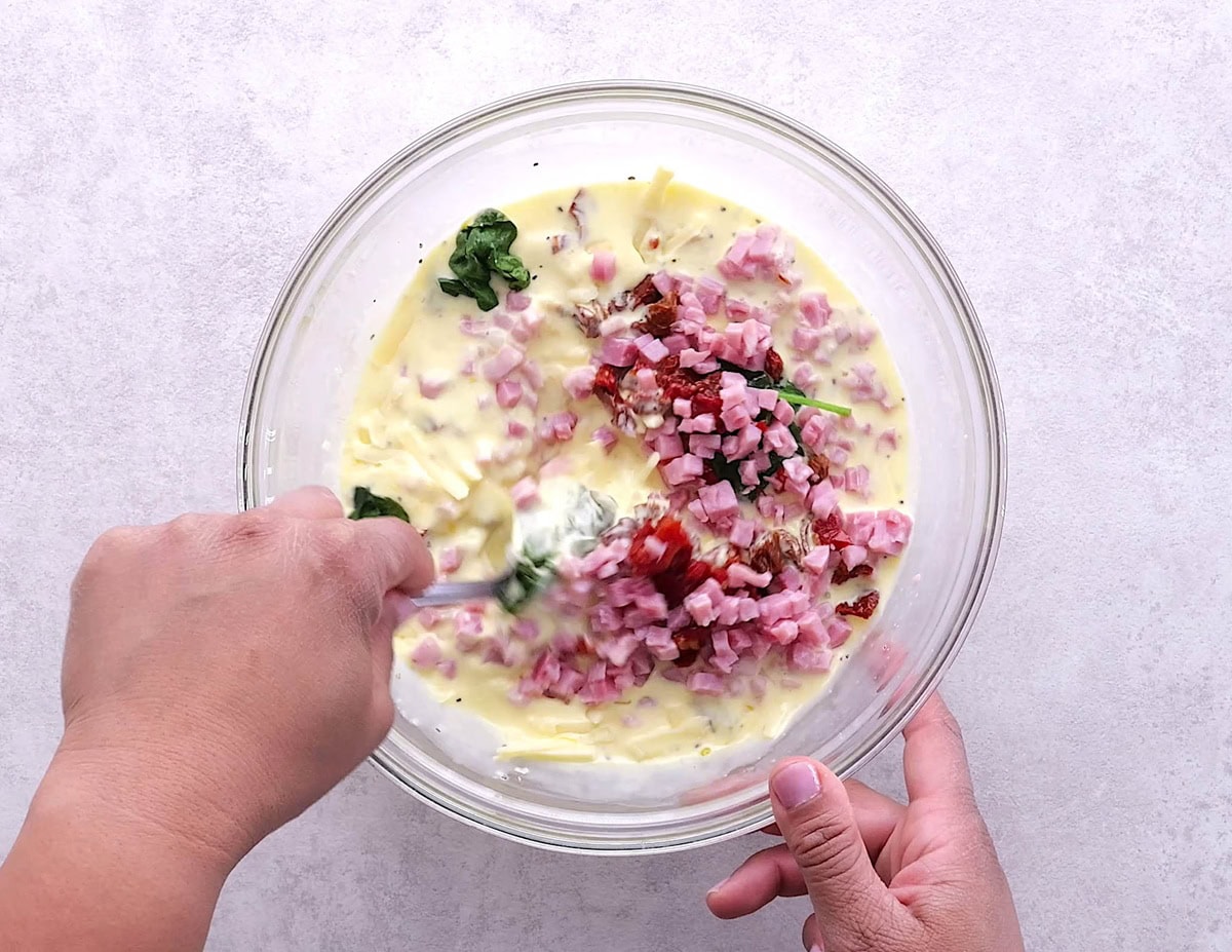 Mixing bowl full of egg custard with spinach, ham, and sun-dried tomatoes being mixed in.