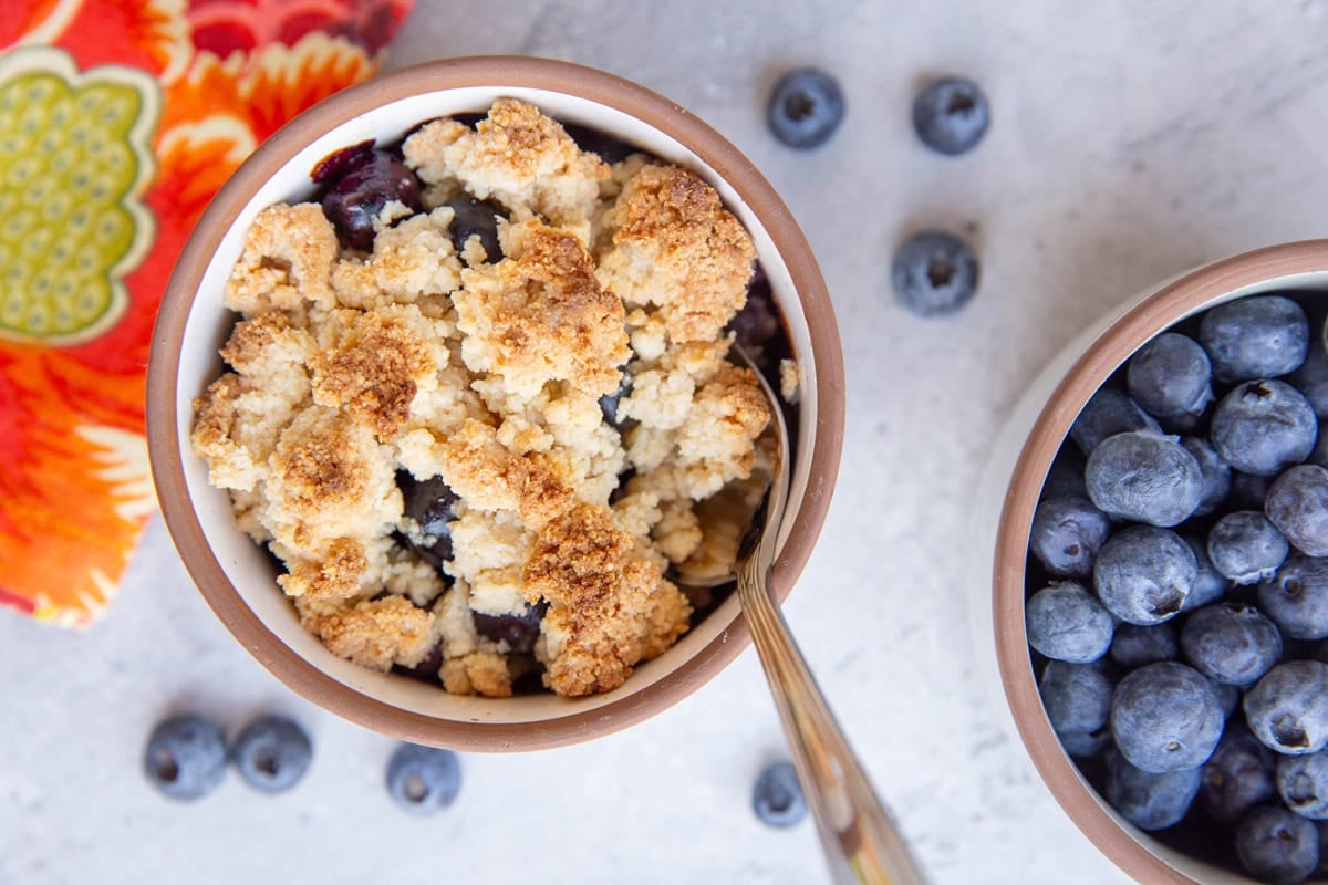 Blueberry crumble in a ramekin with a bowl of fresh berries to the side.