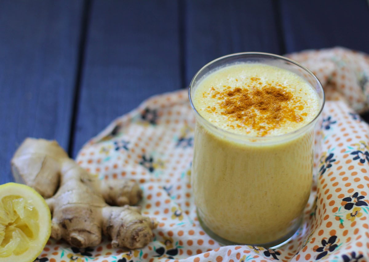 Glass of turmeric lassi on top of a napkin with fresh lemon and ginger to the side.