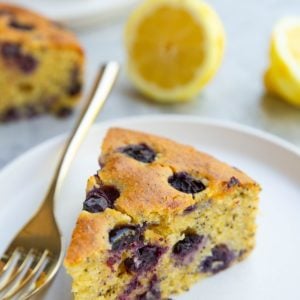 Slice of lemon poppy seed blueberry cake on a plate with a gold fork to the side. Another slice of cake in the background and fresh cut lemons.