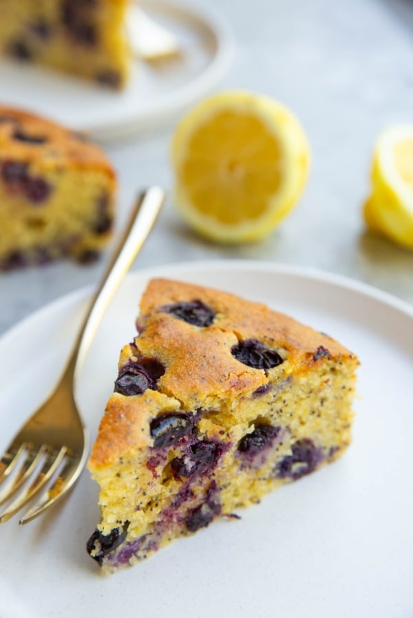Slice of lemon poppy seed blueberry cake on a plate with a gold fork to the side. Another slice of cake in the background and fresh cut lemons.