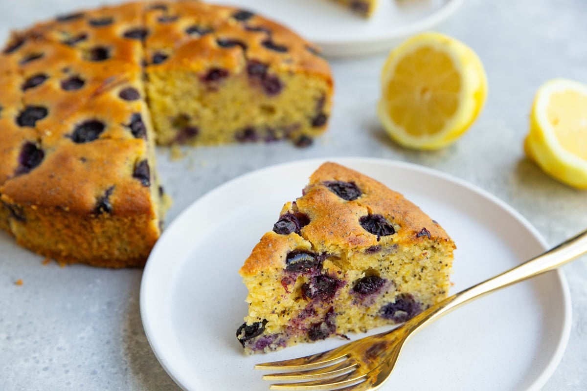 Slice of blueberry lemon cake on a plate with the rest of the cake in the background and cut lemons to the side.