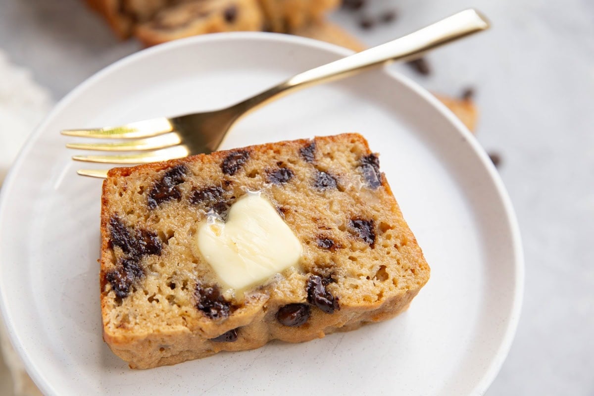 Slice of chocolate chip bread on a plate with butter on top. A golden fork to the side.