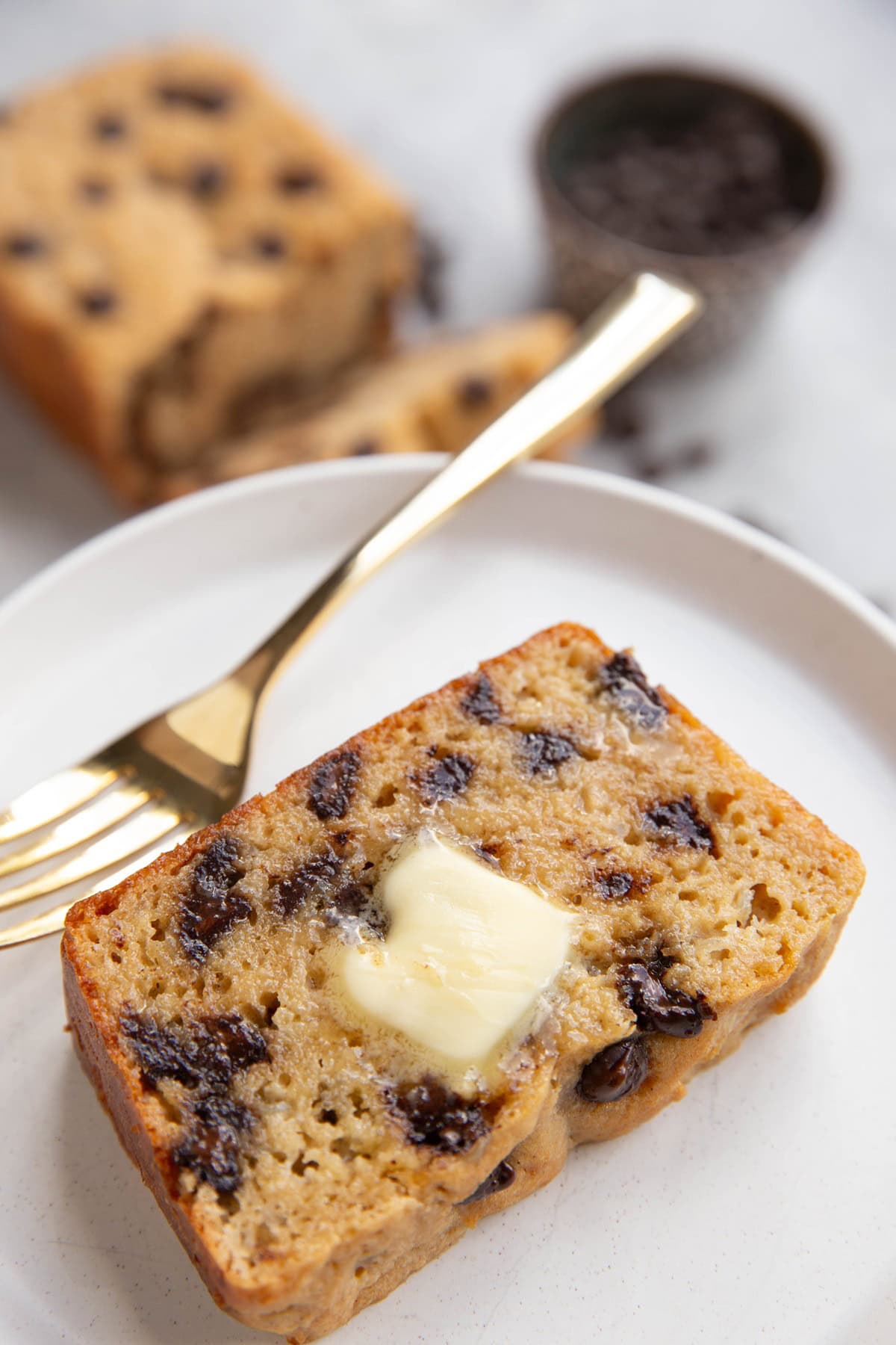 Thick slice of chocolate chip bread on a white plate with melted butter. A gold fork to the side.