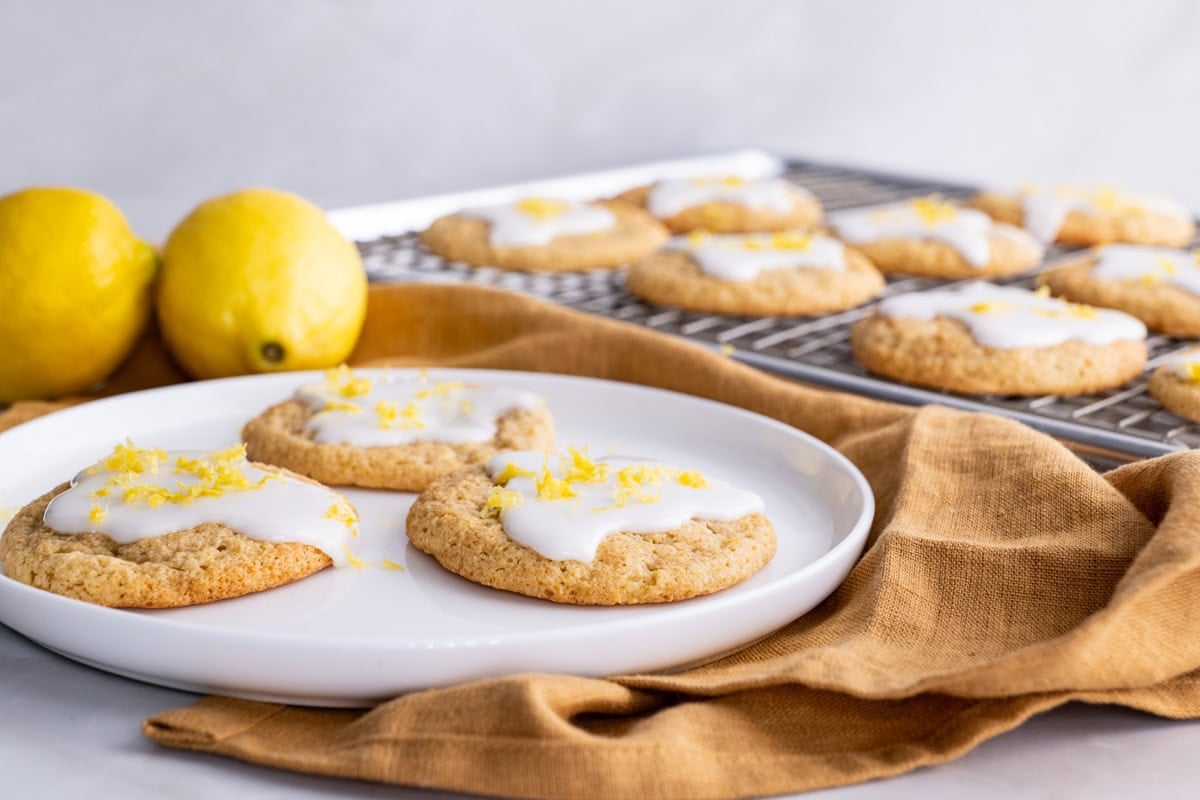 Three almond flour lemon cookies on a plate with lemons in the background and the rest of the cookies on a tray in the background.