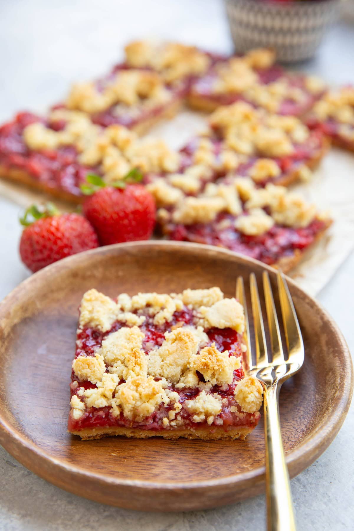 Wooden plate with a slice of strawberry crumb bar on top and the rest of the crumb bars in the background.