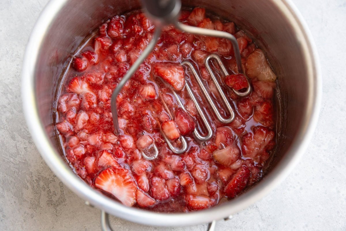 Saucepan full of strawberries being mashed by a potato masher.