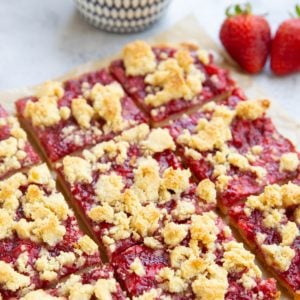Strawberry crumb bars cut into slices on a sheet of parchment paper with a bowl of strawberries in the background.