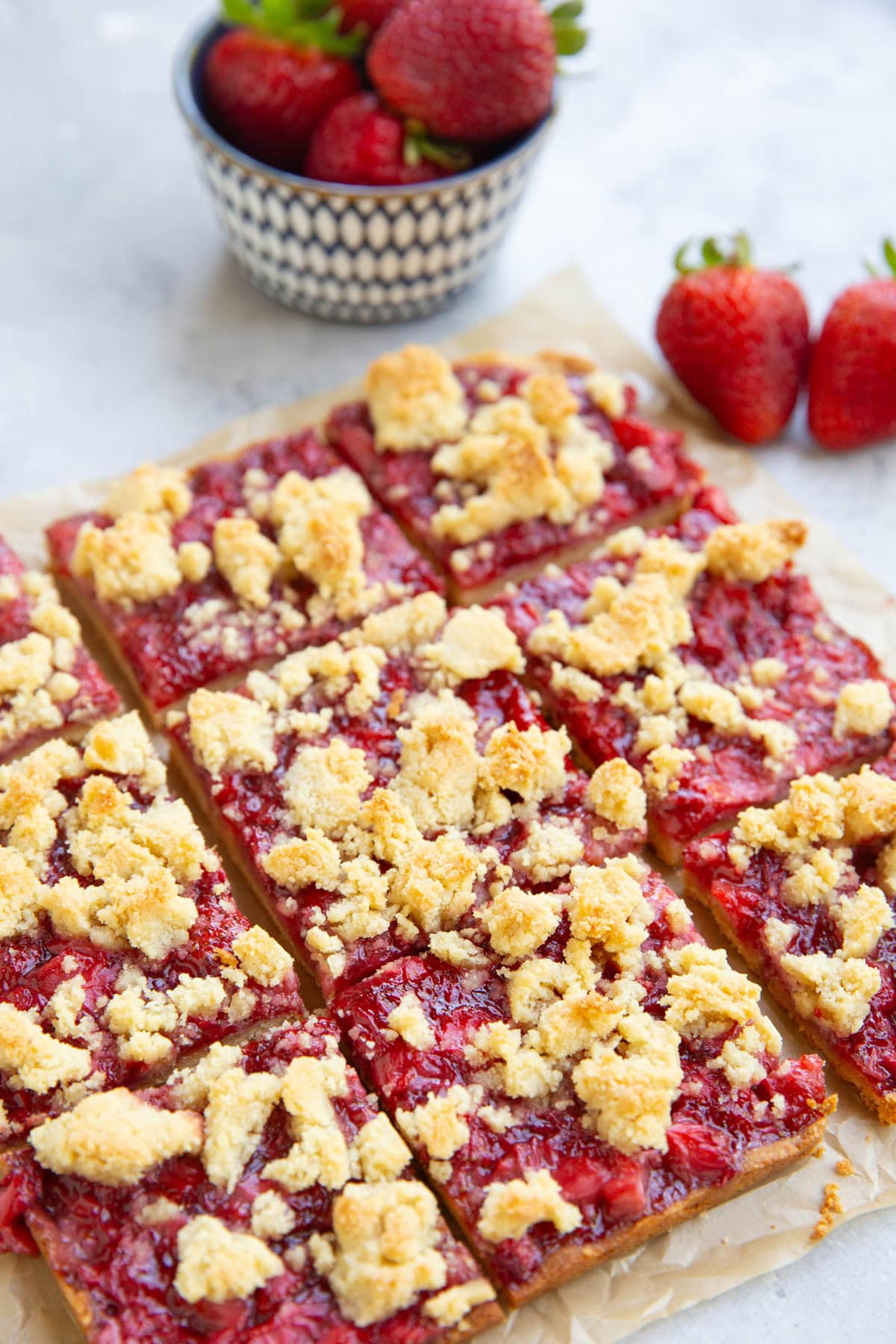 Strawberry crumb bars cut into slices on a sheet of parchment paper with a bowl of strawberries in the background.