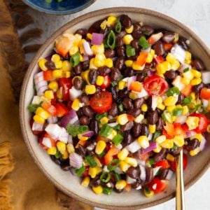 Big bowl of black bean salad with corn, tomato, and onions. A bowl of green onions to the side and a serving spoon.