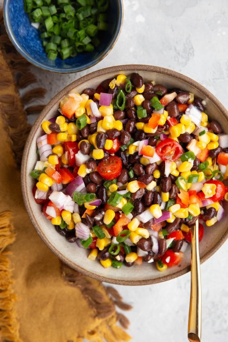 Big bowl of black bean salad with corn, tomato, and onions. A bowl of green onions to the side and a serving spoon.