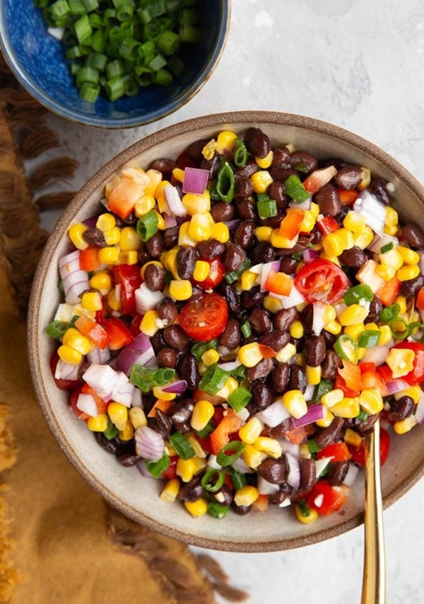 A bowl of colorful black bean salad with corn, cherry tomatoes, red onion, and chopped green onions. A small blue bowl of green onions sits nearby on a textured surface with a brown cloth.
