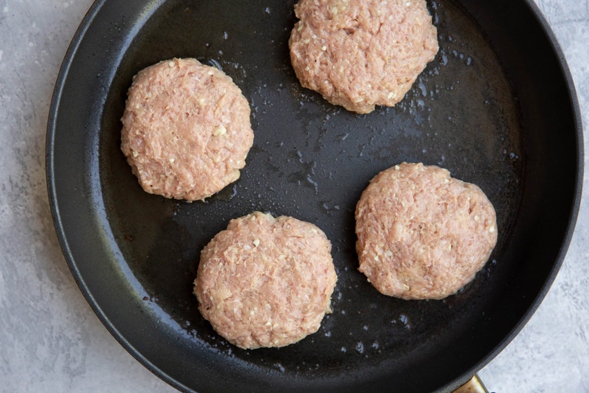Four raw, uncooked chicken patties cooking in a nonstick frying pan.