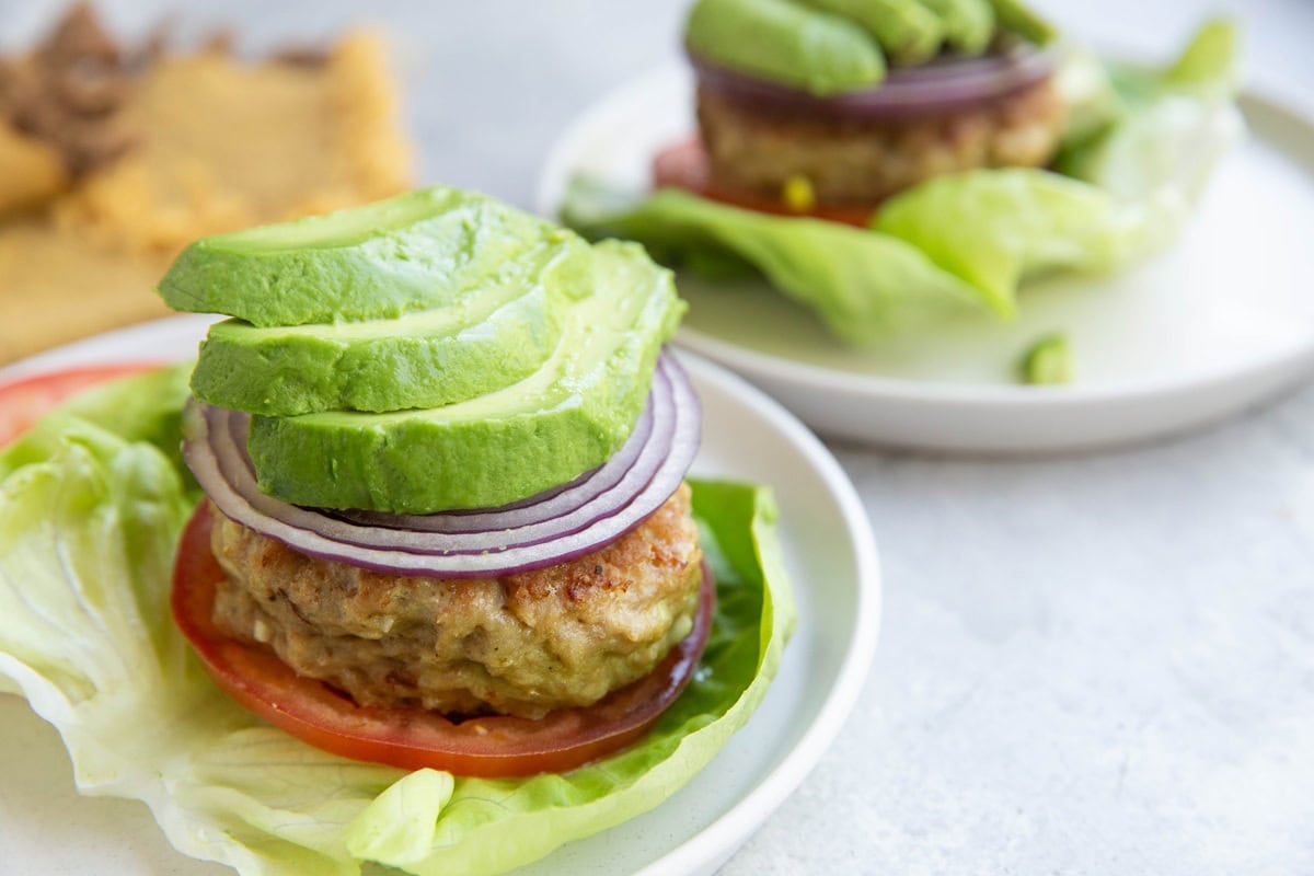 Two lettuce wrapped chicken burgers on plates with onion, tomato, and sliced avocado.