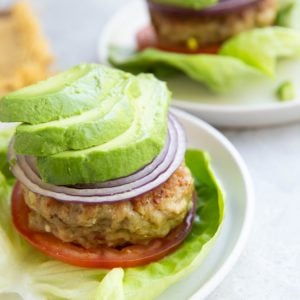 A lettuce-wrapped chicken burger topped with sliced tomato, red onion, and avocado with another chicken patty with toppings in the background.
