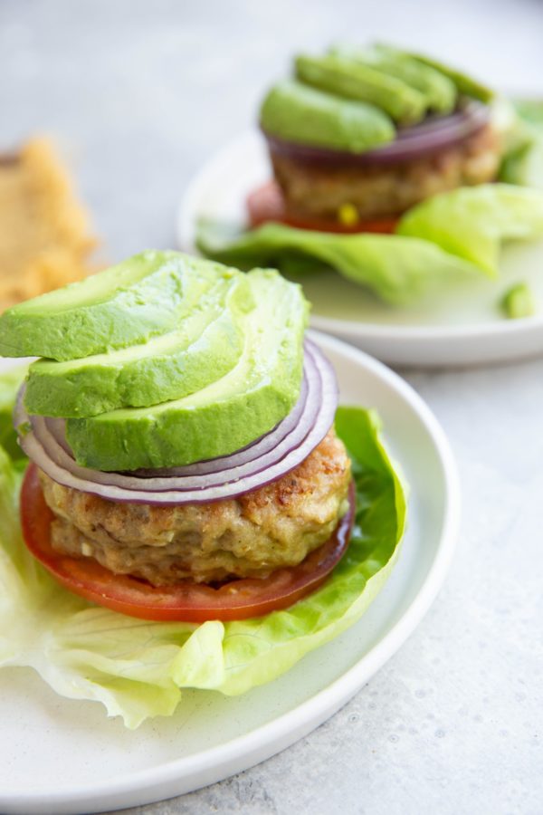 A lettuce-wrapped chicken burger topped with sliced tomato, red onion, and avocado with another chicken patty with toppings in the background.