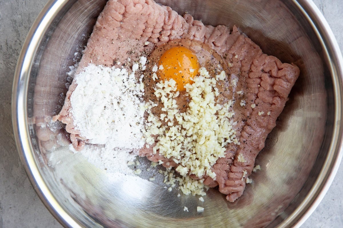 A metal bowl containing ground chicken, a raw egg yolk, minced garlic, flour, black pepper, and other seasonings, ready to be mixed together.